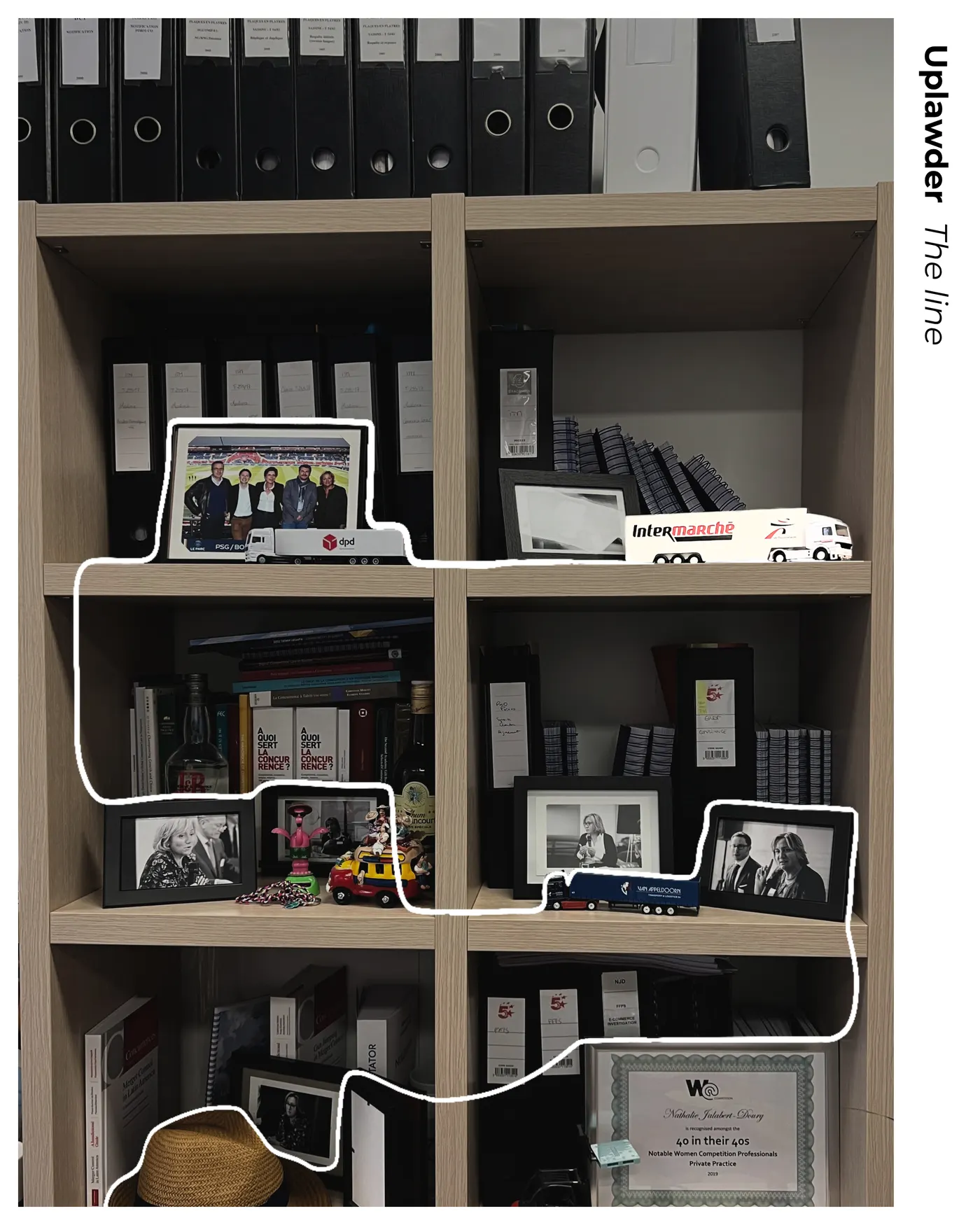 La bibliothèque du bureau, traversée par le trait blanc de la série : cartons de dossiers, camions miniatures Intermarché, DPD et Van Appeldoorn, ouvrages de droit de la concurrence et certificat 40 in their 40s.